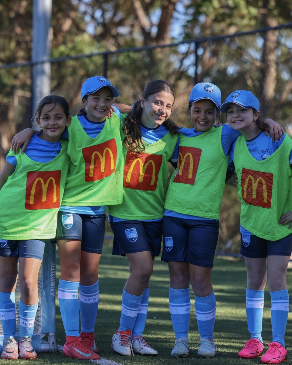 Five young girls in Sydney FC kit and training bibs smiling arm in arm on the pitch