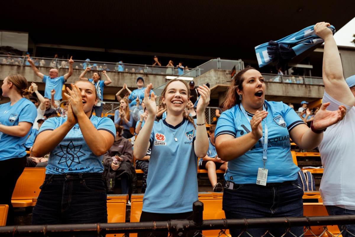Sydney FC fans in sky blue jerseys cheering and clapping in the stands